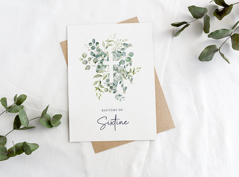 Faire-part de baptême avec une croix blanche sur du feuillage d'eucalyptus