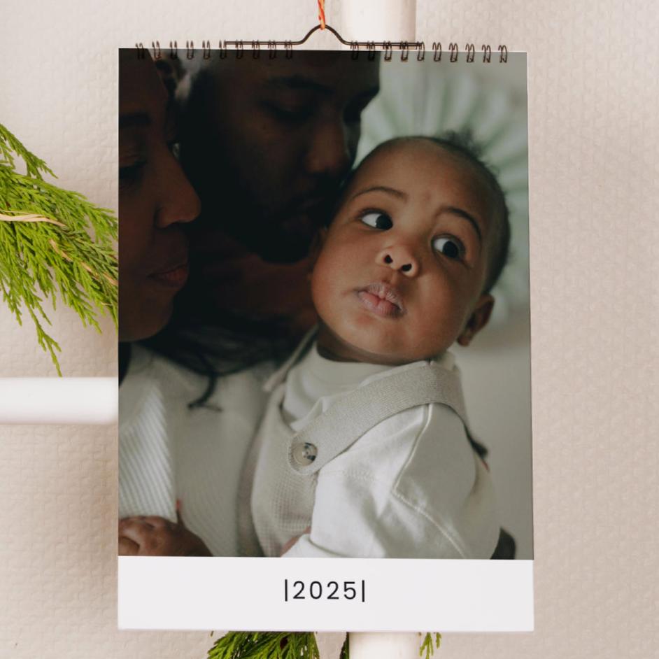 A3-Wandkalender im Hochformat, der an einer Wand hängt und auf der Titelseite geöffnet ist, die ein großes Foto eines Babys mit dem Text „2025“ darunter zeigt.