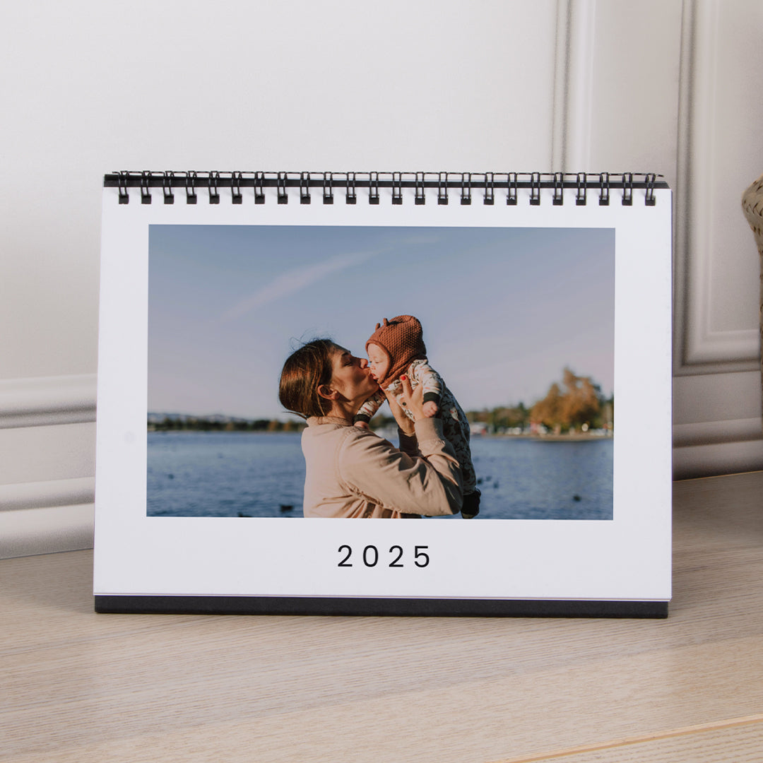 Tischkalender im quadratischen Format, der auf einem Tisch steht und auf dem Deckblatt mit dem Foto einer Mutter mit Baby und dem Text „2025“ darunter aufgeschlagen ist.